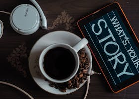 Flat lay of coffee cup, headphones, and tablet with 'What's Your Story' on screen.